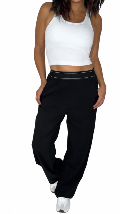 A woman modeling a pair of black wide-leg sweatpants with a thin white stripe detail along the waistband, paired with a white crop top and white sneakers, against a plain white background.