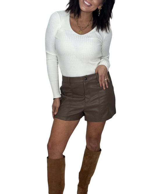 Woman wearing a cream v-neck top with brown leather shorts and brown boots on a white background