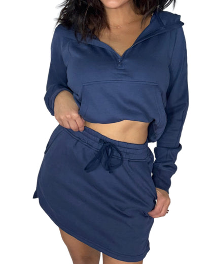 Close profile of a woman wearing a matching navy blue loungewear set , half-zip hoodie and a drawstring mini skort, shown against a plain white background