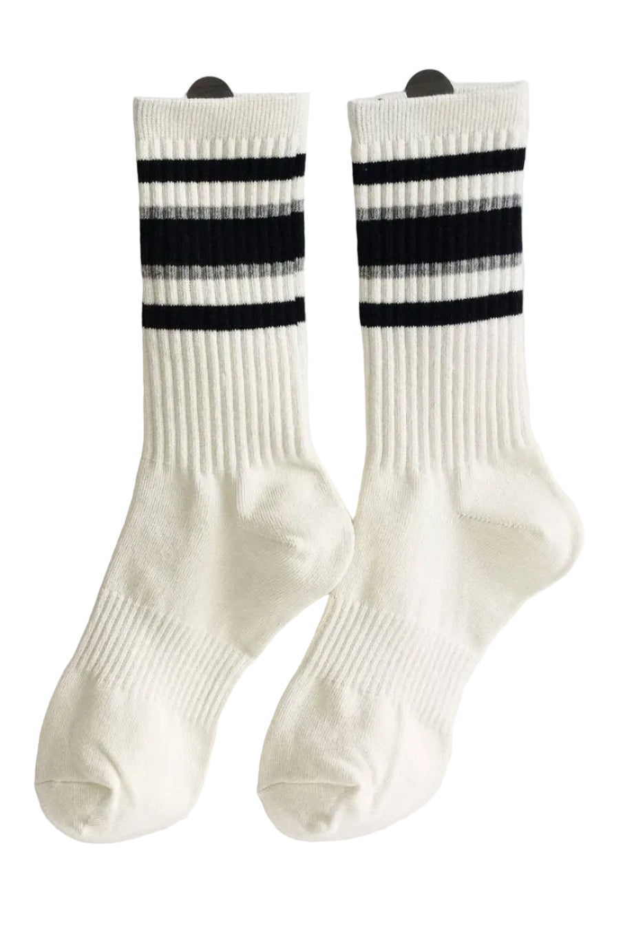 A pair of off-white ribbed crew socks with three black horizontal stripes near the cuff, displayed on a white wall 