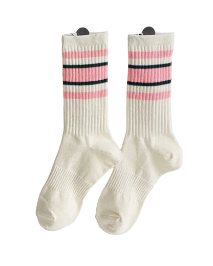 A pair of ribbed crew socks in off-white with pink and black horizontal stripes near the cuff, displayed on a white wall n